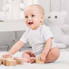 Uśmiechnięte niemowlę w białym body bawi się drewnianymi klockami na dywanie.