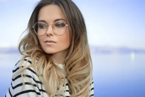 Blondynka w okularach o okrągłych oprawkach, w bluzie w paski, na tle błękitnego nieba i wody.