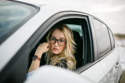 Kobieta w okularach za kierownicą białego samochodu. Zastanawia się, jakie okulary do jazdy samochodem wybrać, by zapewnić sobie komfort i bezpieczeństwo.