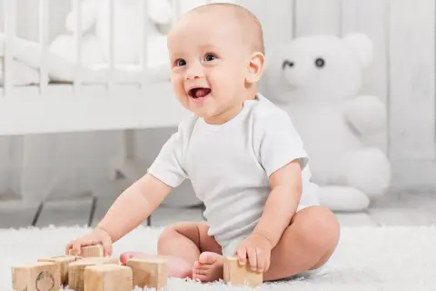 Uśmiechnięte niemowlę w białym body bawi się drewnianymi klockami na dywanie.