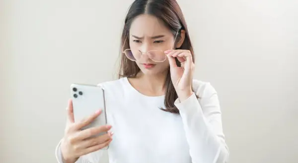 Kobieta w okularach z trudem patrzy na ekran telefonu.