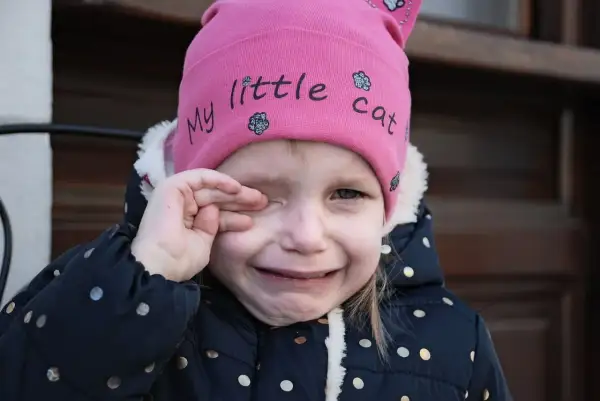 Mała dziewczynka w różowej czapce z napisem "my little cat" płacze, pocierając oko. Wygląda, jakby coś jej wpadło, może opiłek w oku.