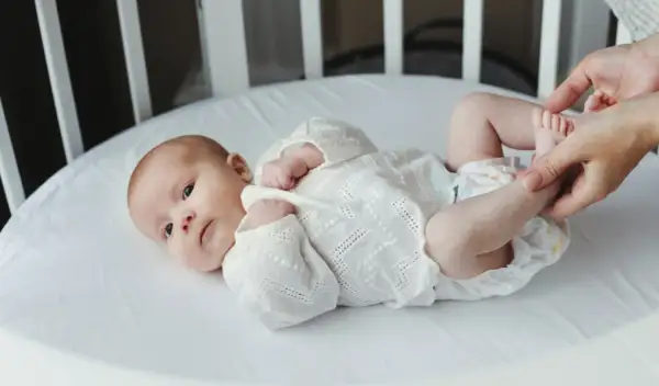 Noworodek w białej koszulce leży w łóżeczku, a ręce rodzica dotykają jego stóp.